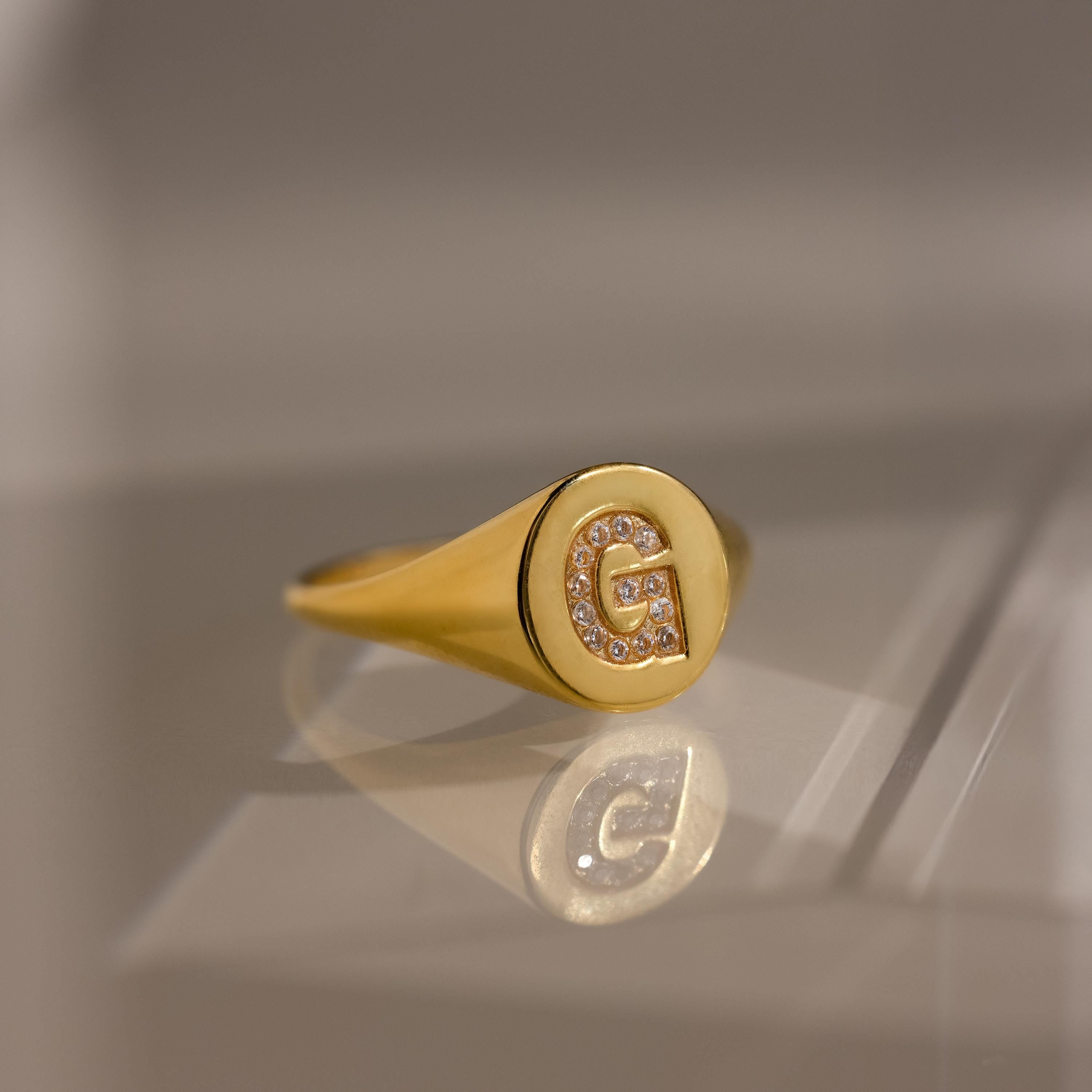 The Pave Initial Signet Ring in 18K Gold features a crystal-studded letter G engraved on its round face, reflecting light beautifully—an eye-catching statement piece.