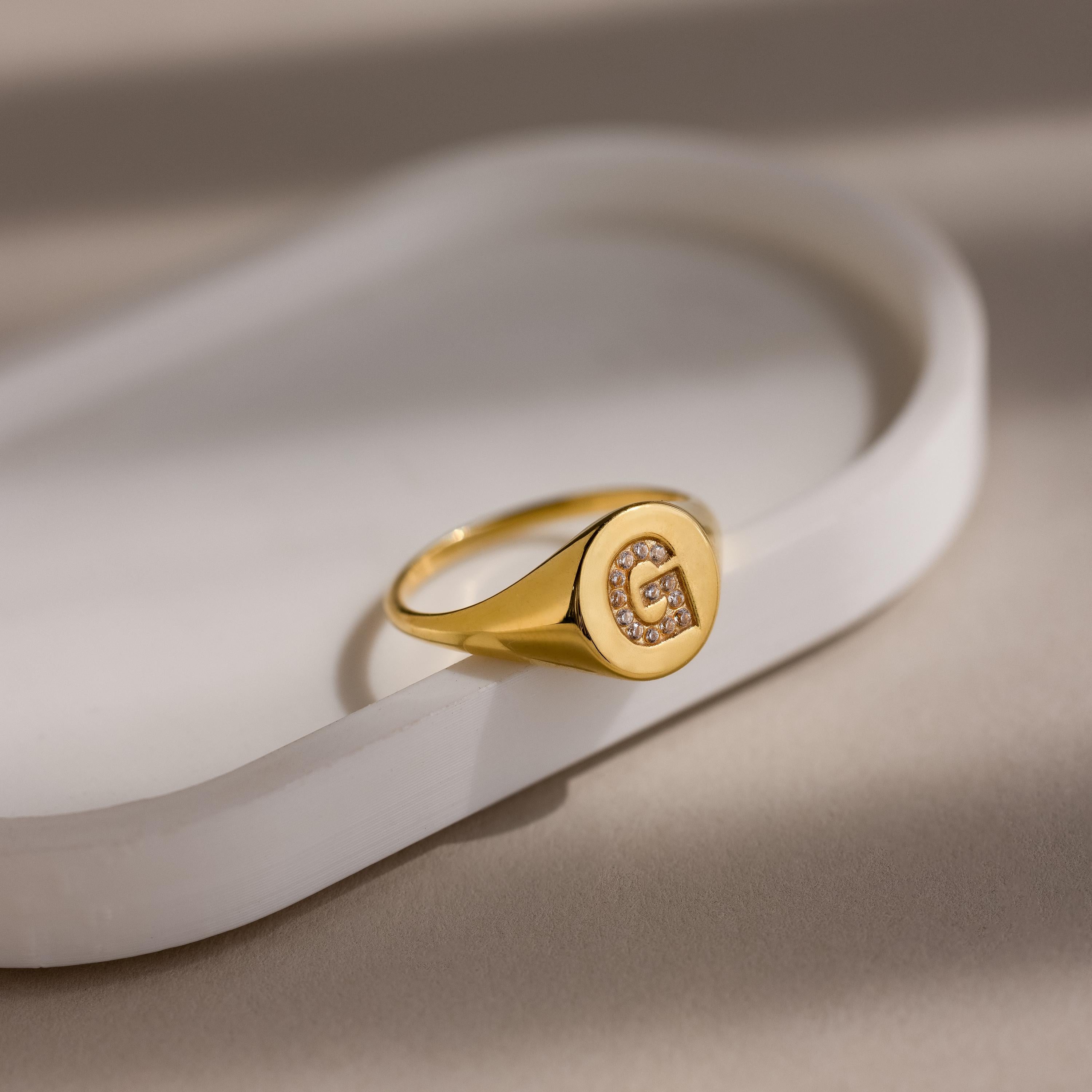 The Pave Initial Signet Ring in 18K Gold features a crystal-studded letter G, beautifully showcased on a white oval tray—perfect for making a subtle yet elegant statement.