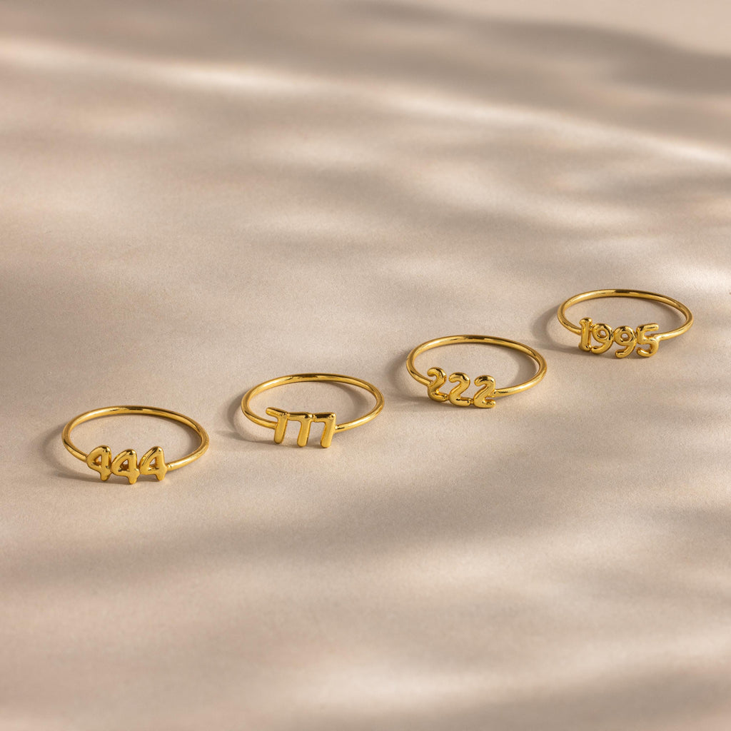 Four gold Bubble Numbers Rings with “944,” “777,” “222,” and “1995” are arranged on a beige background.