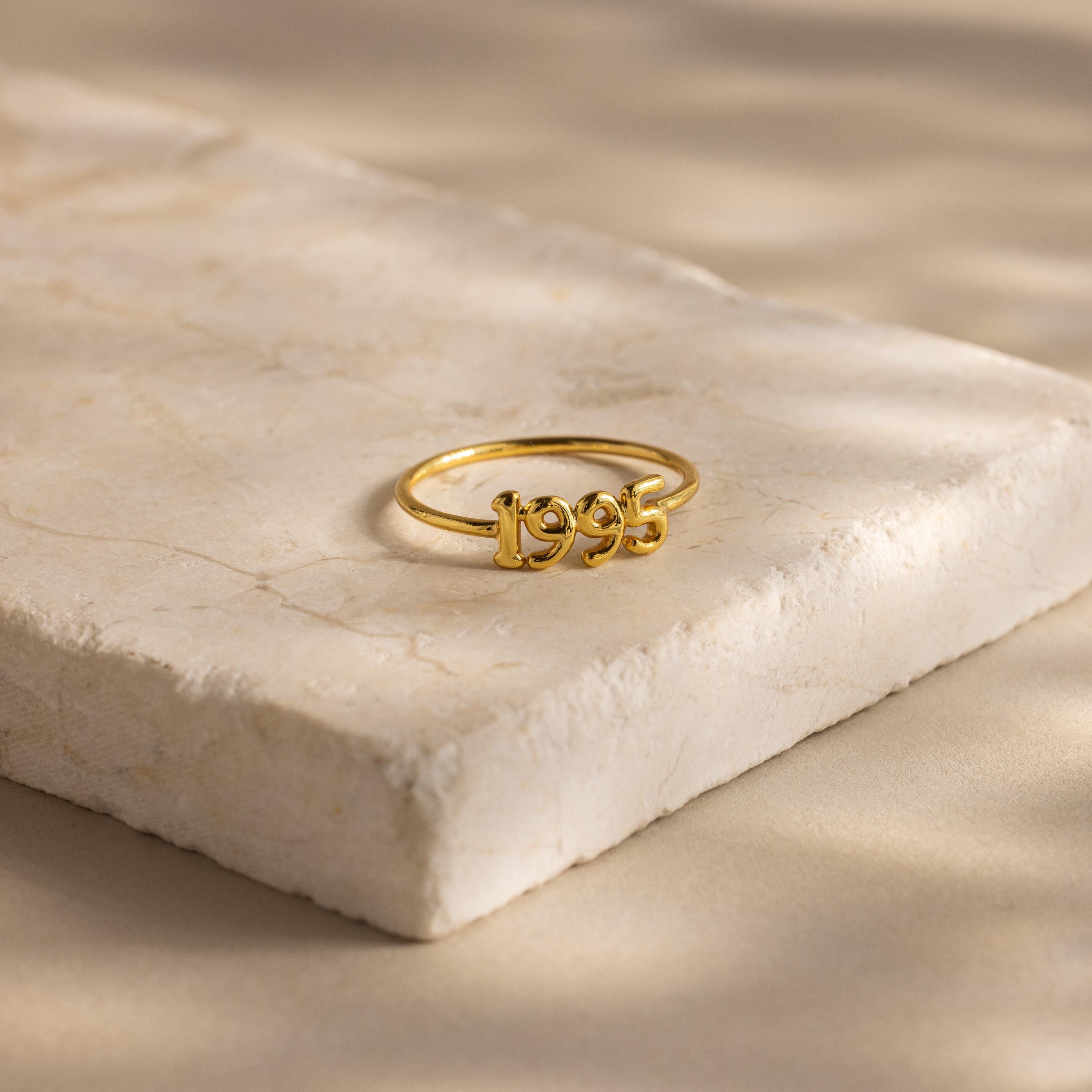 A gold ring featuring the year “1995” is displayed on a cream stone platform in soft lighting.