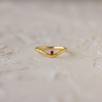 The Amethyst Drop Curve Ring features a delicate gold band with a small amethyst centerpiece, beautifully displayed on a textured light surface. Perfect as a stacking ring for an elegant and subtle accent.