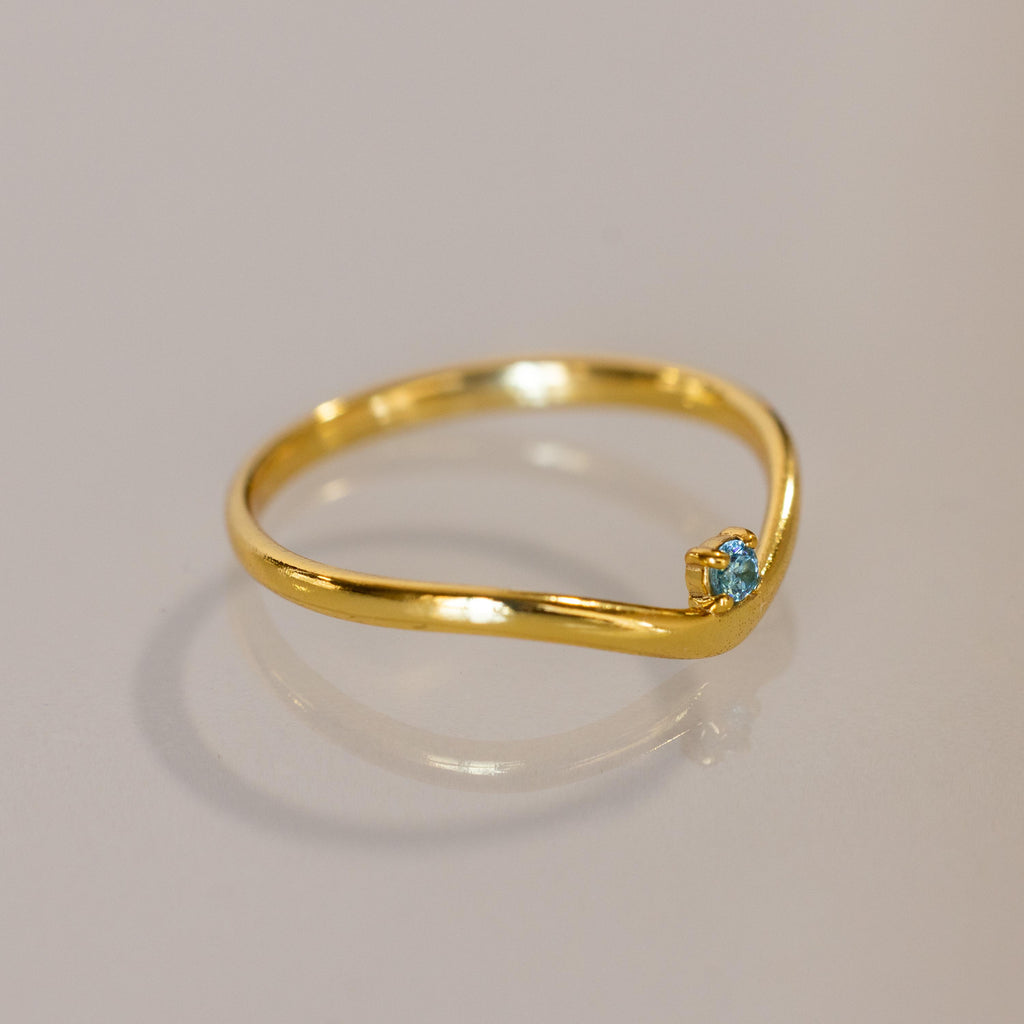 The Blue Zircon Drop Curve Ring features a thin gold band with a small blue zircon gemstone on a gentle curve, ideal as a delicate stacking ring and shown on a light surface.