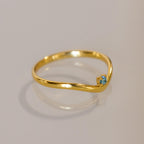 The Blue Zircon Drop Curve Ring features a thin gold band with a small blue zircon gemstone on a gentle curve, ideal as a delicate stacking ring and shown on a light surface.