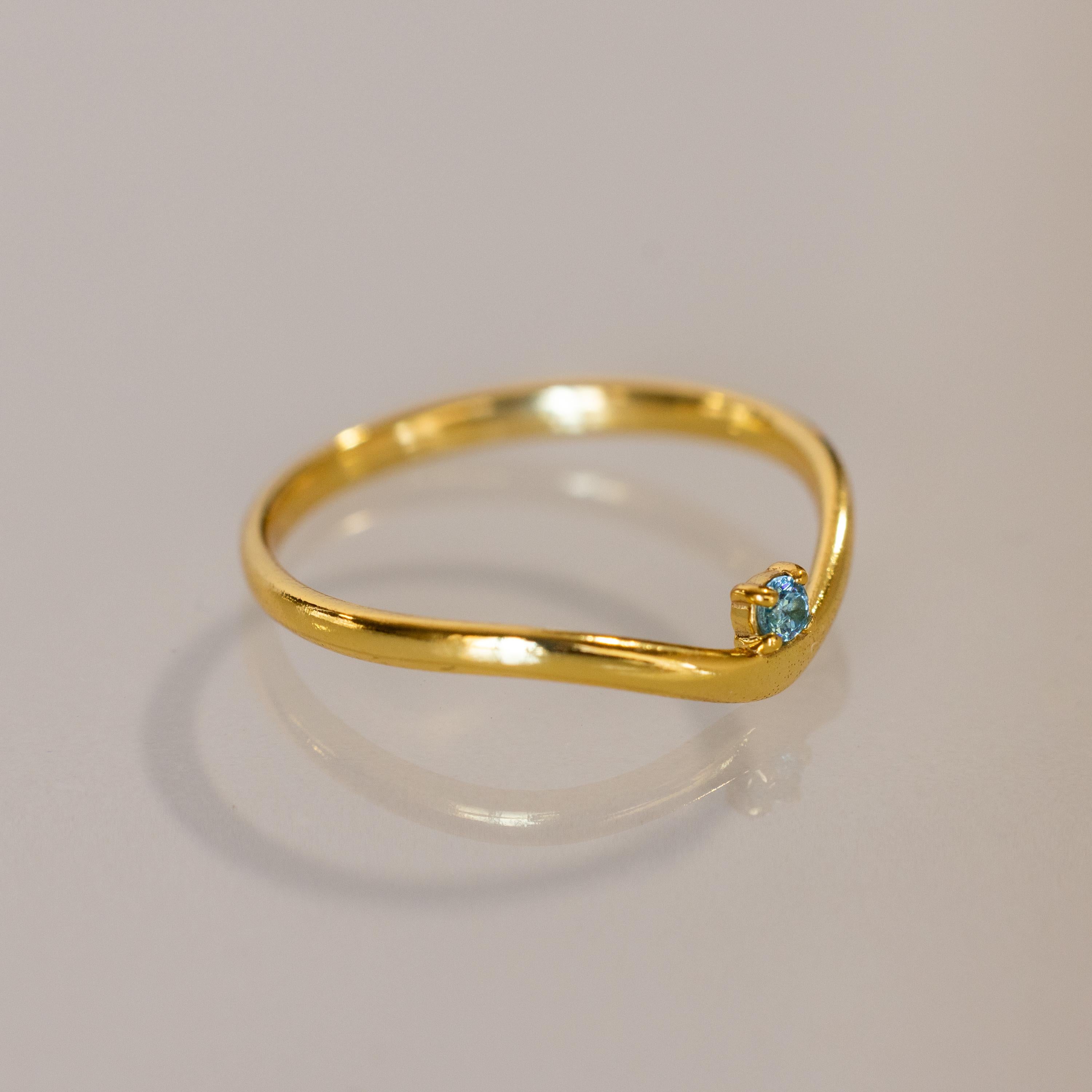 The Blue Zircon Drop Curve Ring features a thin gold band with a small blue zircon gemstone on a gentle curve, ideal as a delicate stacking ring and shown on a light surface.