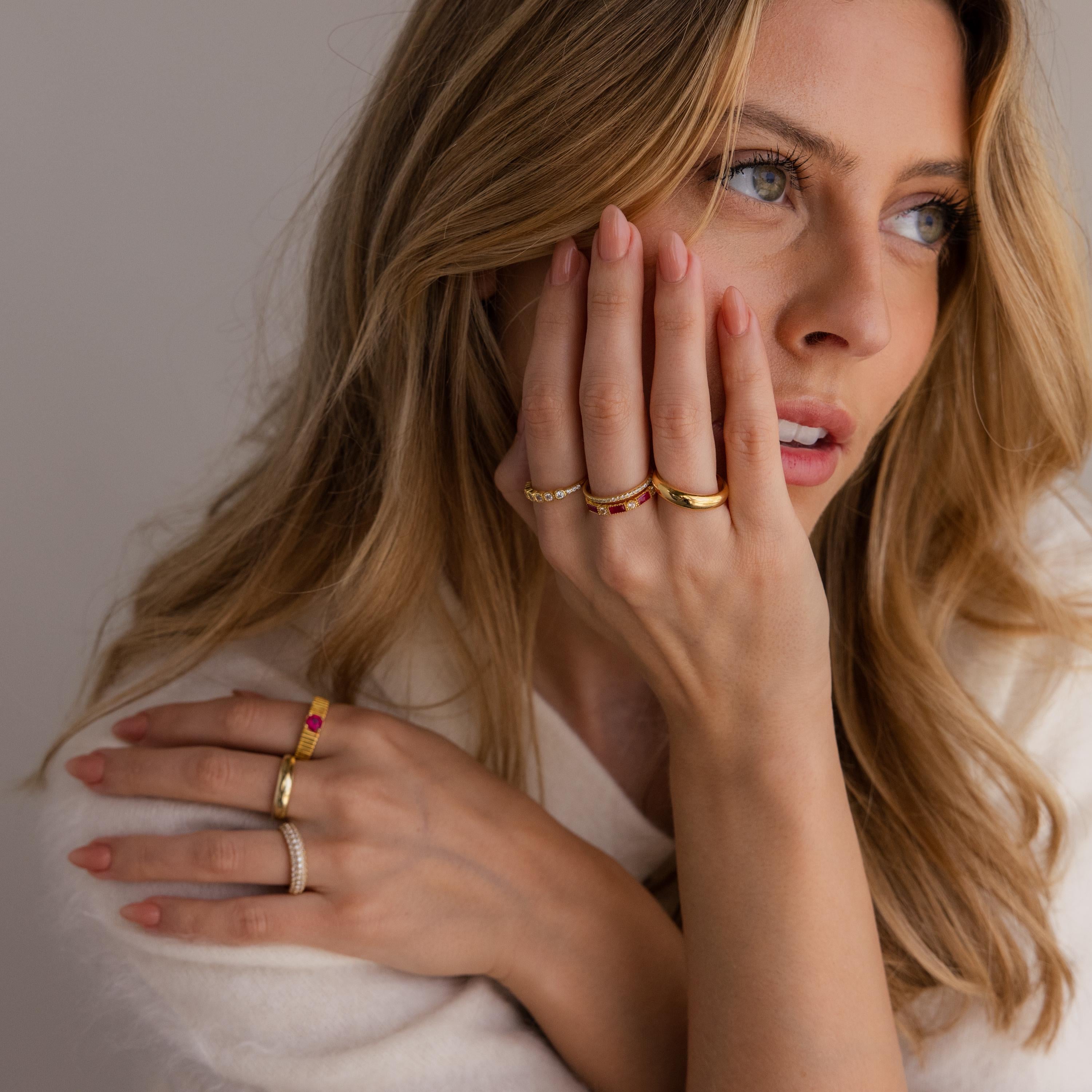A woman with long blonde hair, wearing a white sweater and the Mixed Art Deco Birthstone Ring, touches her face and looks to the side, gold rings accenting her hand.