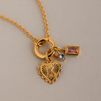 The Cable Circle Charm Necklace features a gold chain with a heart pendant, blue gem, and rectangular pink stone—an elegant statement piece shown against a beige background.