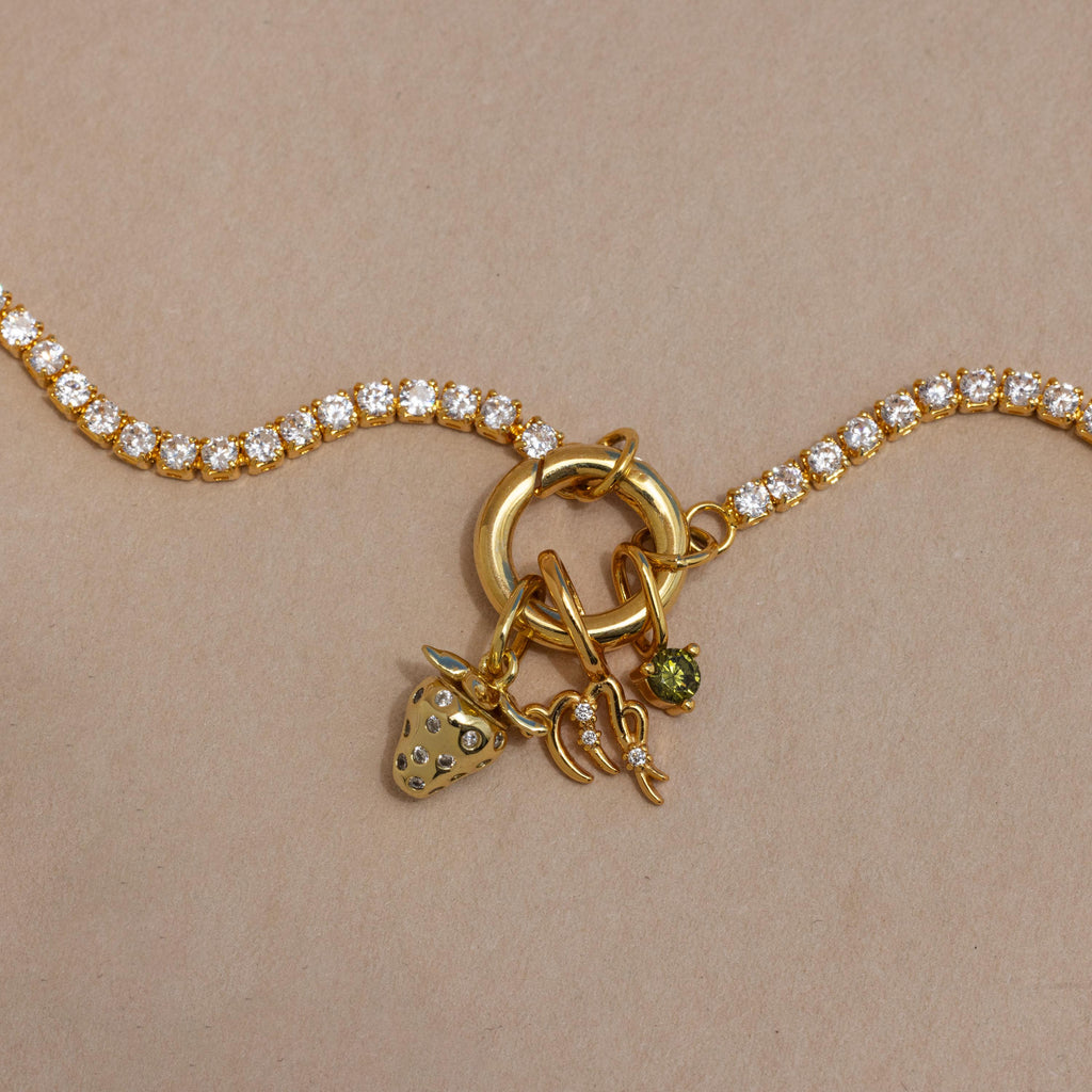 The Diamond Tennis Clasp Necklace features a gold chain with clear round gemstones and three gold charms—a strawberry, 1997 number, and green stone—secured with a rolo clasp. Shown on a light brown background, its perfect for wedding jewelry.