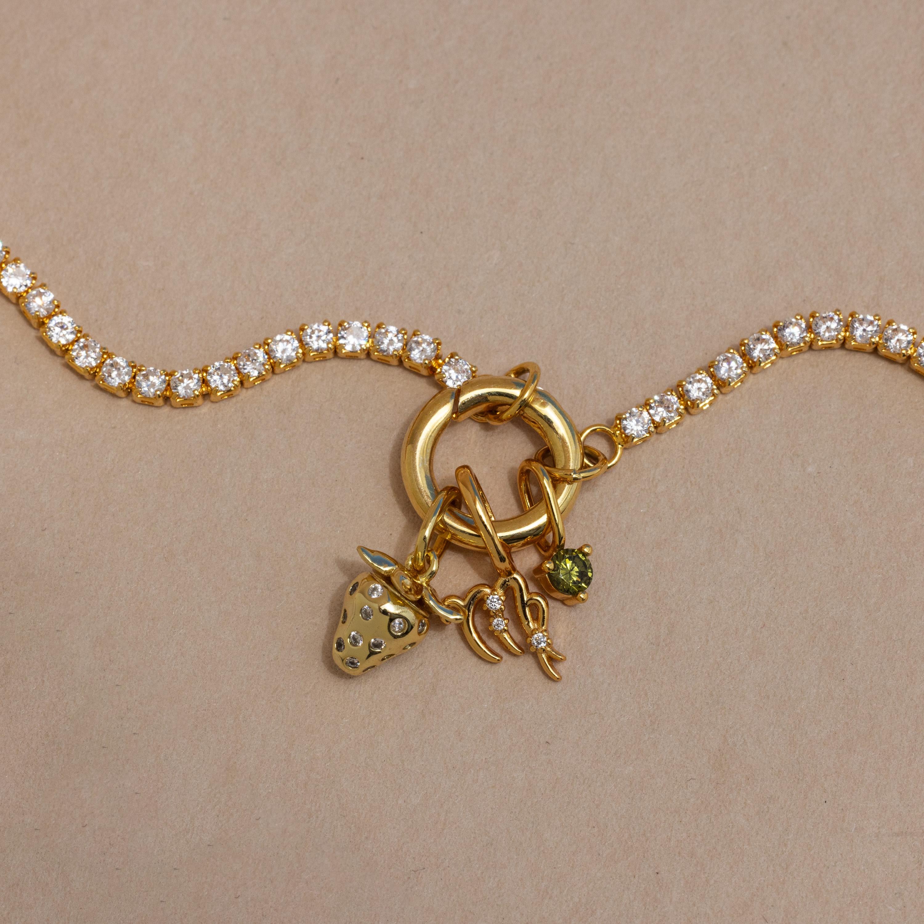 The Diamond Tennis Clasp Necklace features a gold chain with clear round gemstones and three gold charms—a strawberry, 1997 number, and green stone—secured with a rolo clasp. Shown on a light brown background, its perfect for wedding jewelry.