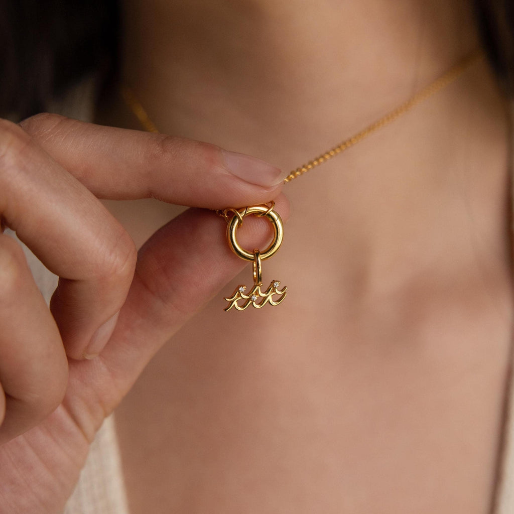 A close-up of a hand holding a gold Aquarius charm pendant on a circular bail, hanging from a dainty gold chain.