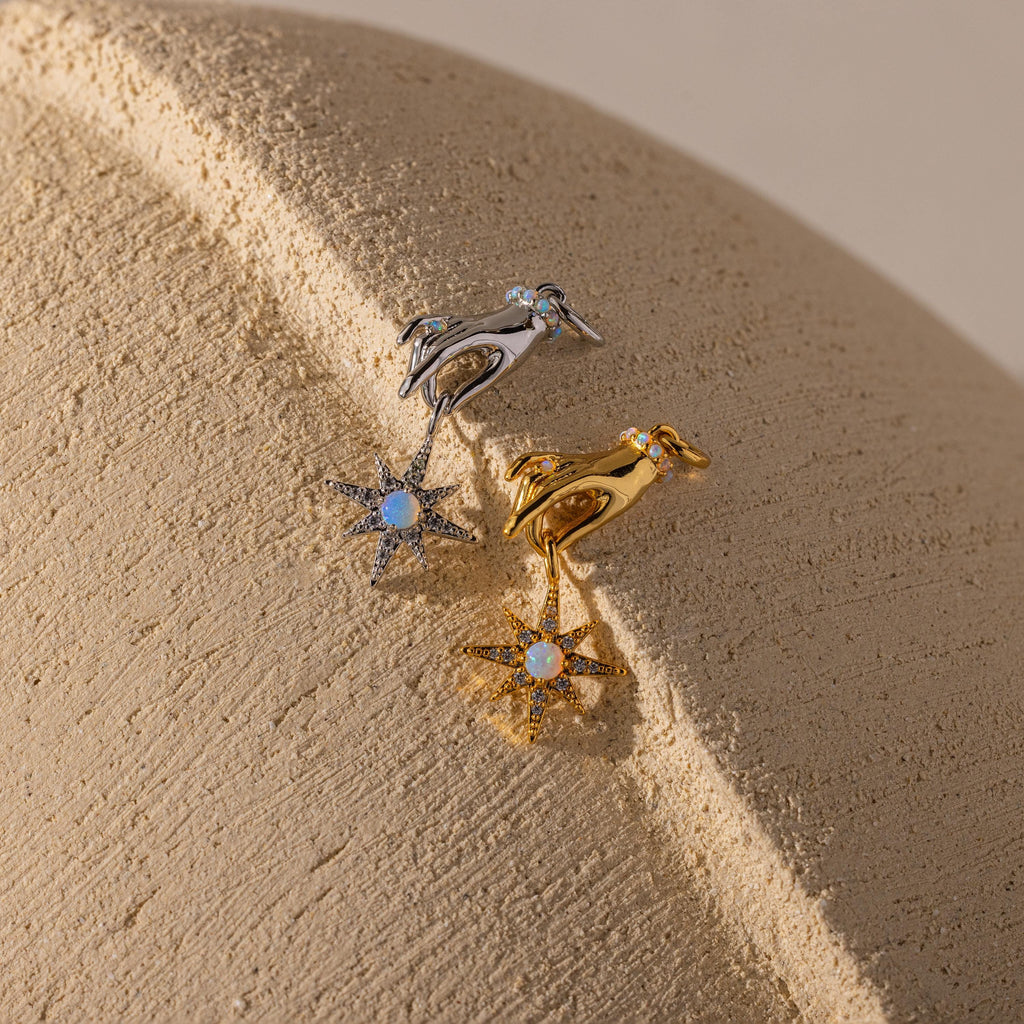 Two hand-and-star pendants—one in silver, one in gold—are styled side-by-side on a textured neutral surface.