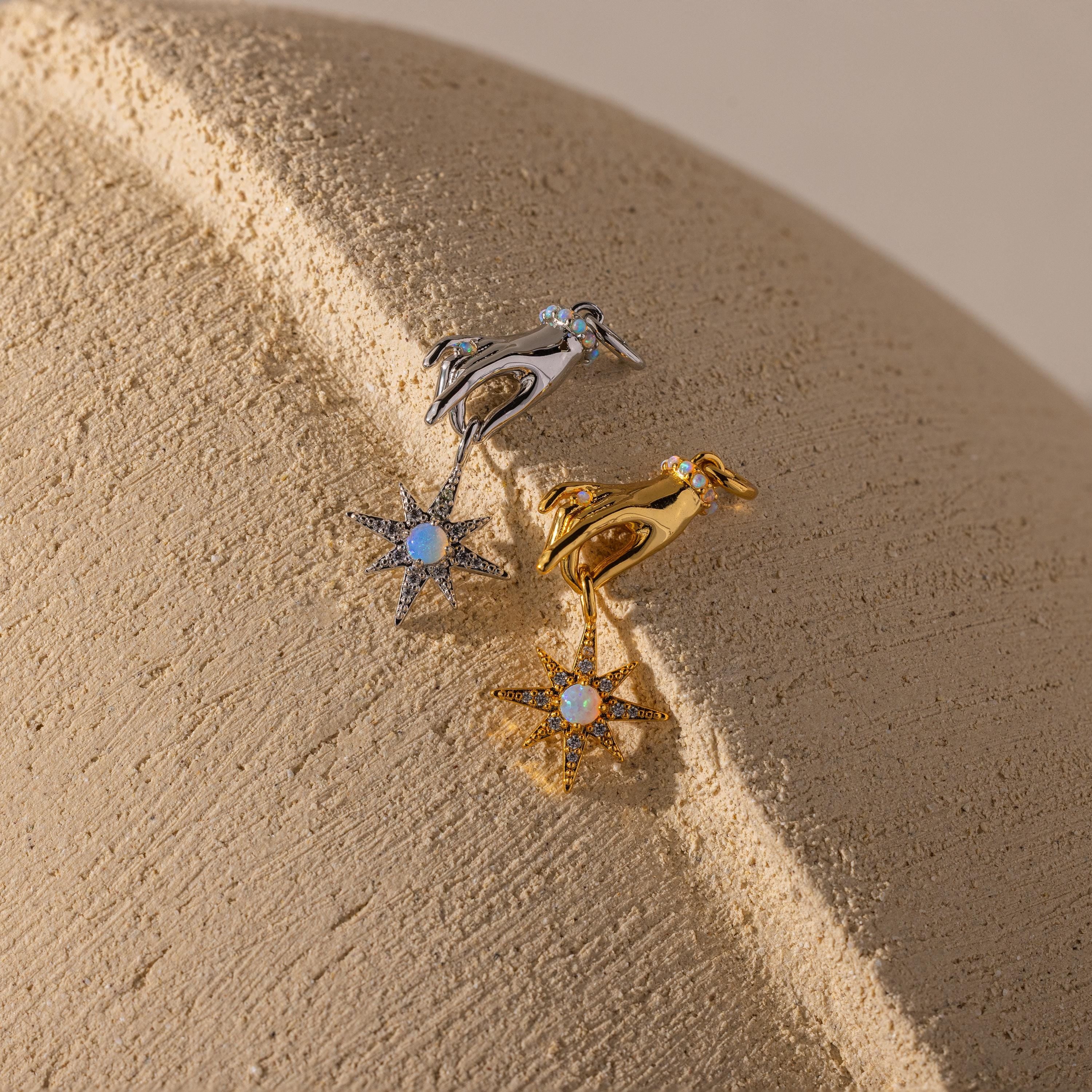 Two hand-and-star pendants—one in silver, one in gold—are styled side-by-side on a textured neutral surface.