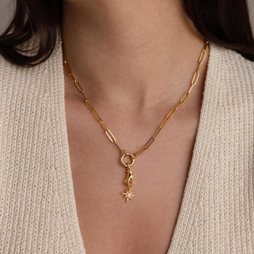 The gold hand and star pendant hangs from a chunky gold paperclip chain necklace worn by a model