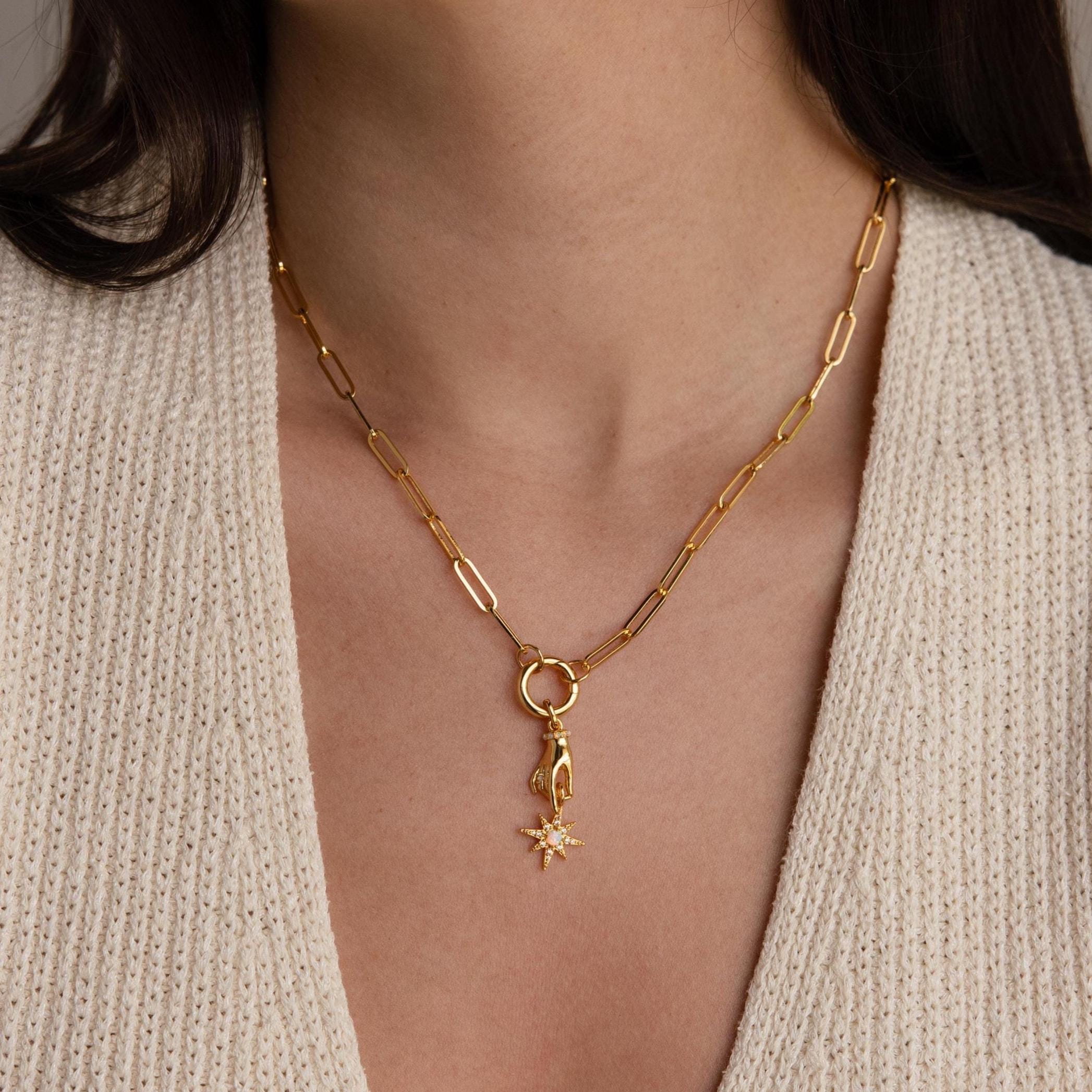 The gold hand and star pendant hangs from a chunky gold paperclip chain necklace worn by a model