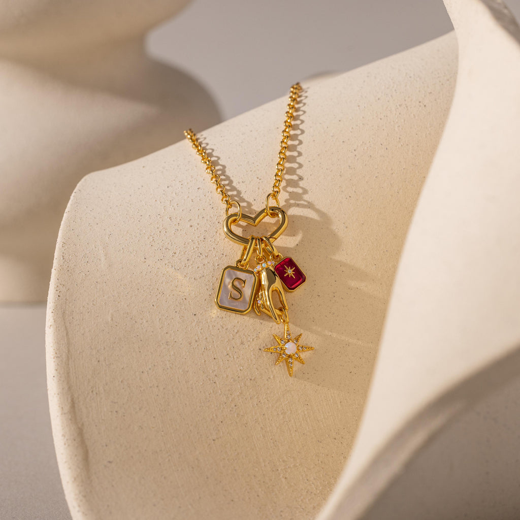 Gold necklace with heart link and four charms: letter "S", red tag with star, wishbone, and starburst with crystals, displayed on a cream bust.
