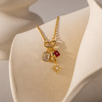 Gold necklace with heart link and four charms: letter "S", red tag with star, wishbone, and starburst with crystals, displayed on a cream bust.