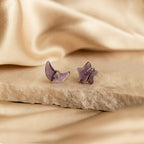 Close up of a pair of purple amethyst moon and star-shaped earrings in silver resting on a beige stone surface.