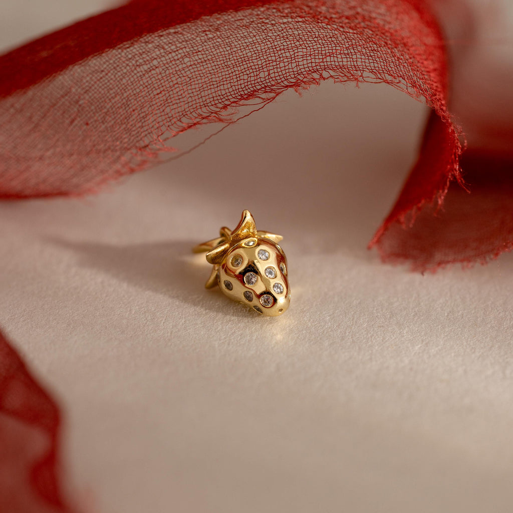 A close-up of a gold strawberry charm with crystal accents, lying on a flat surface partially wrapped in red tulle fabric.