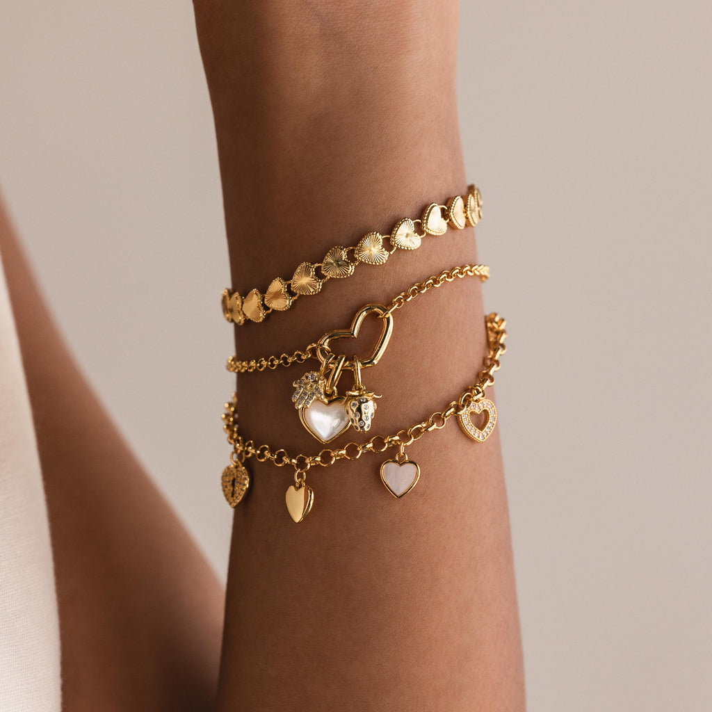 A wrist stacked with multiple gold bracelets. The top is a ribbed heart bracelet, the middle is a chain with a heart-shaped connector and hanging charms, and the bottom one is a link bracelet with multiple heart charms.