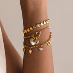 A wrist stacked with multiple gold bracelets. The top is a ribbed heart bracelet, the middle is a chain with a heart-shaped connector and hanging charms, and the bottom one is a link bracelet with multiple heart charms.