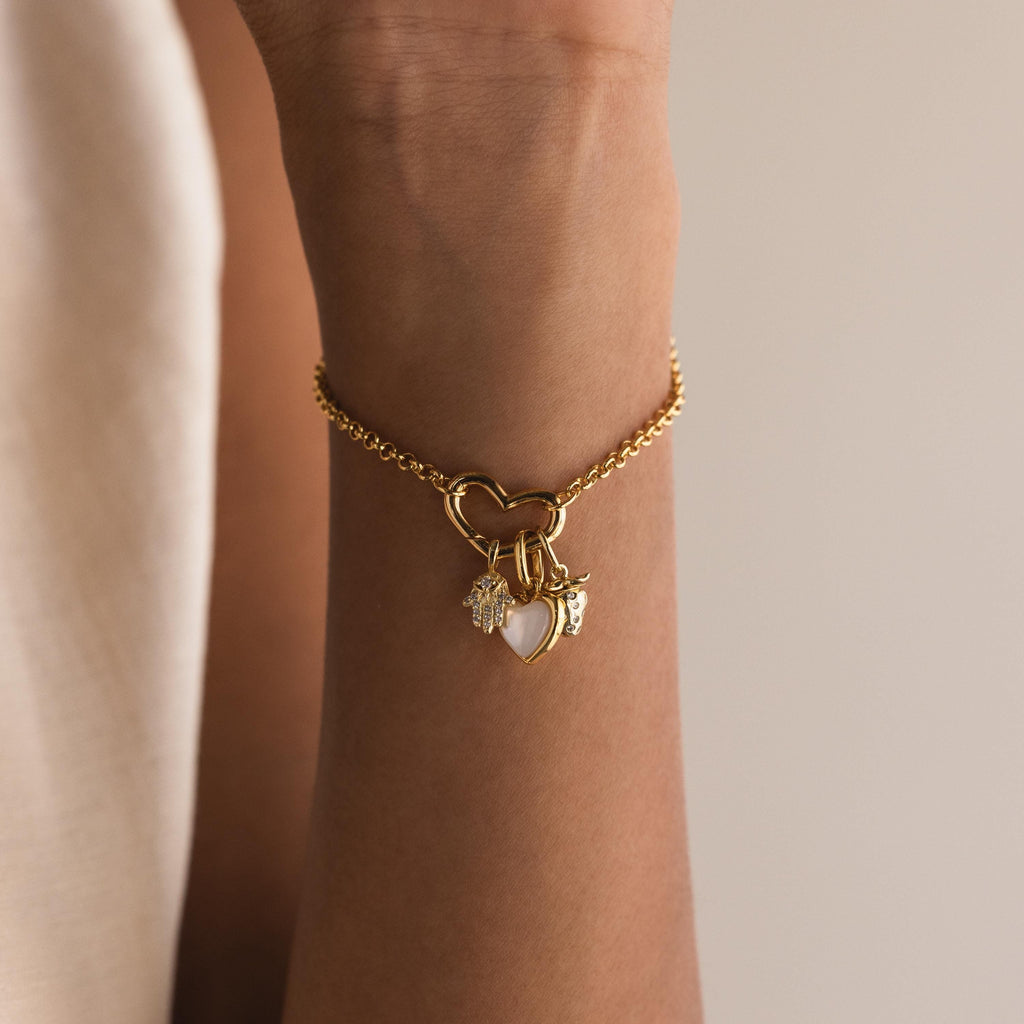 A wrist adorned with a gold bracelet featuring a heart-shaped connector and three hanging charms: a pavé hamsa hand, a mother-of-pearl heart, and the gold strawberry charm.