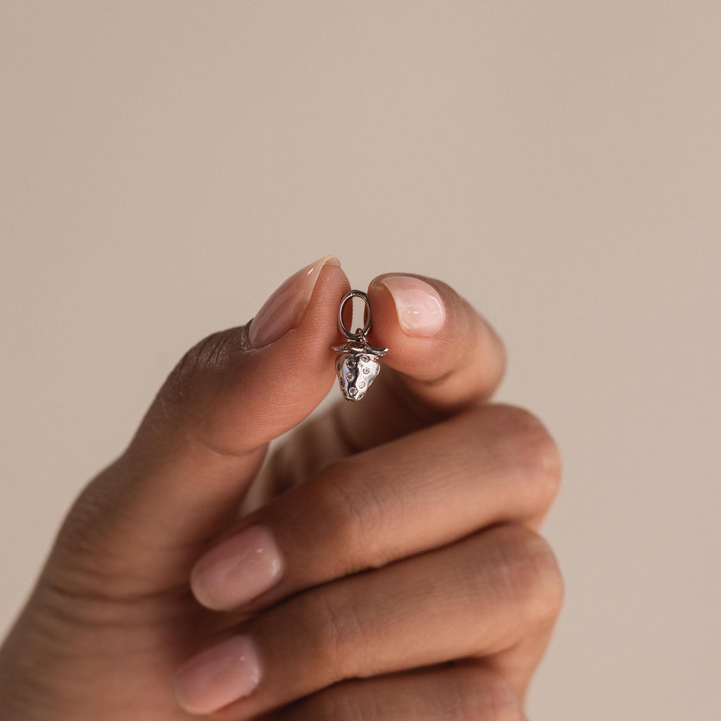 A hand holds a small silver strawberry charm between thumb and index finger, showcasing its miniature size and crystal-studded details.