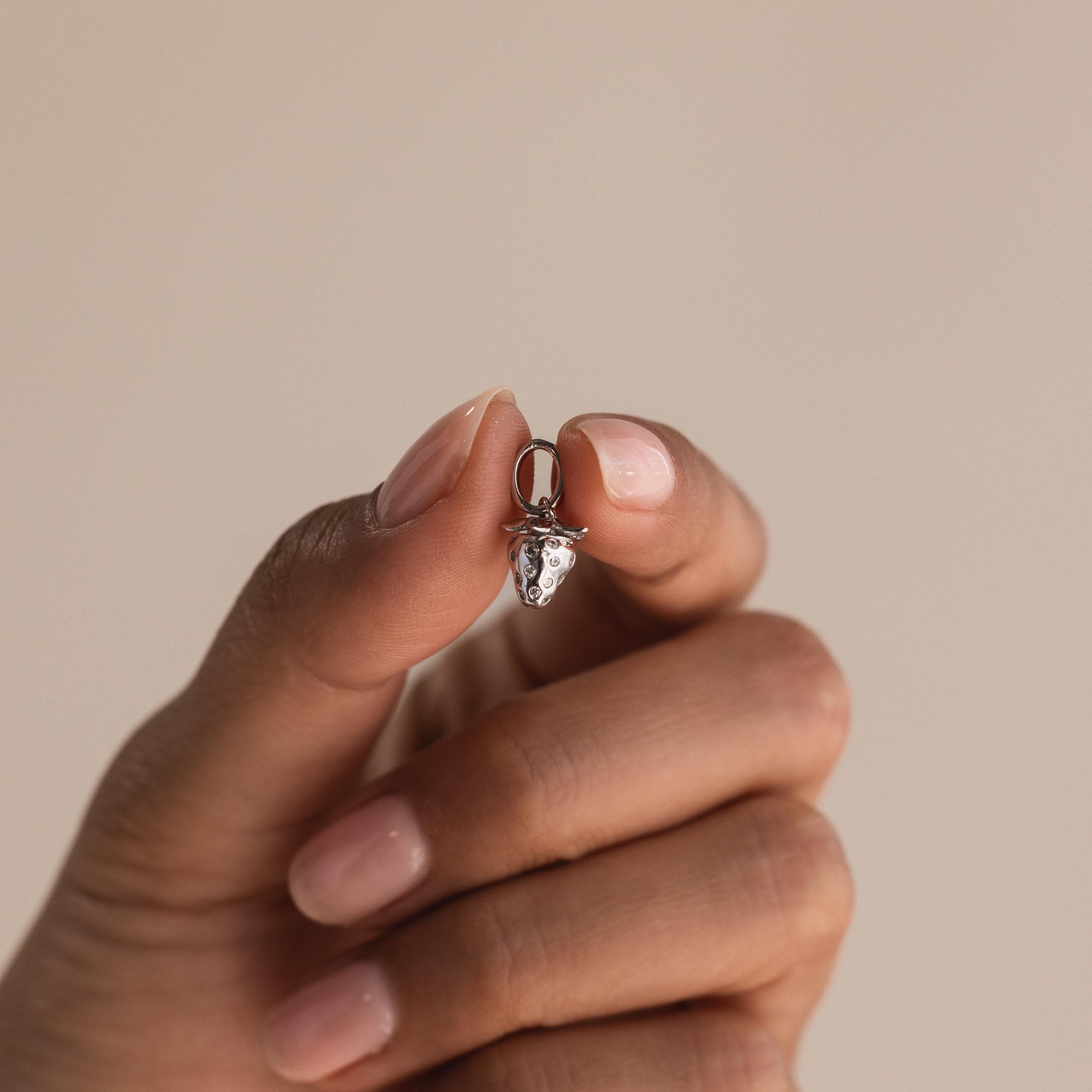 A hand holds a small silver strawberry charm between thumb and index finger, showcasing its miniature size and crystal-studded details.