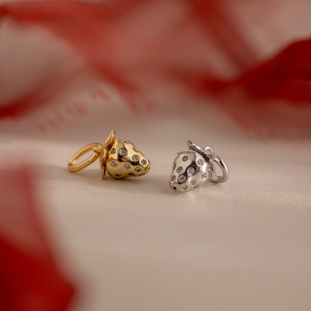 A pair of strawberry-shaped charms, one in gold and one in silver, both embedded with tiny clear crystals, rest side by side on a soft cream surface with red fabric shadows in the background.
