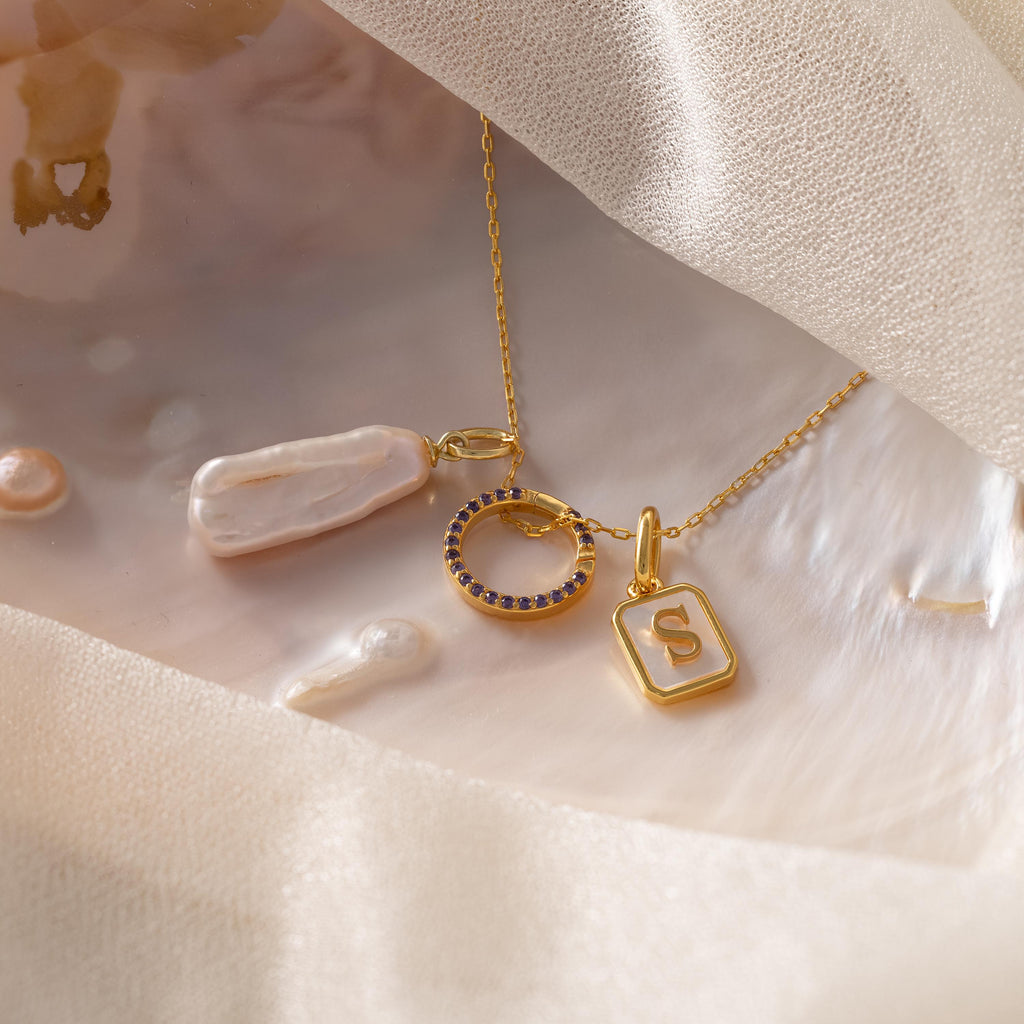 The Pave Amethyst Charm Clip is shown on silky fabric and shells, alongside a gold necklace with pearl, blue gem circle, and initial S charms—featuring a customizable birthstone charm for personalized jewelry.