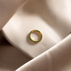 The Pave Emerald Charm Clip, a gold hoop earring with sparkling green gemstones, is perfect for holding charms or adding a birthstone ring charm, shown resting on soft, folded beige fabric.