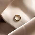 The Pave Sapphire Charm Clip, a gold circular earring adorned with blue gemstones, is perfect as a birthstone ring charm and rests elegantly on smooth beige satin fabric.