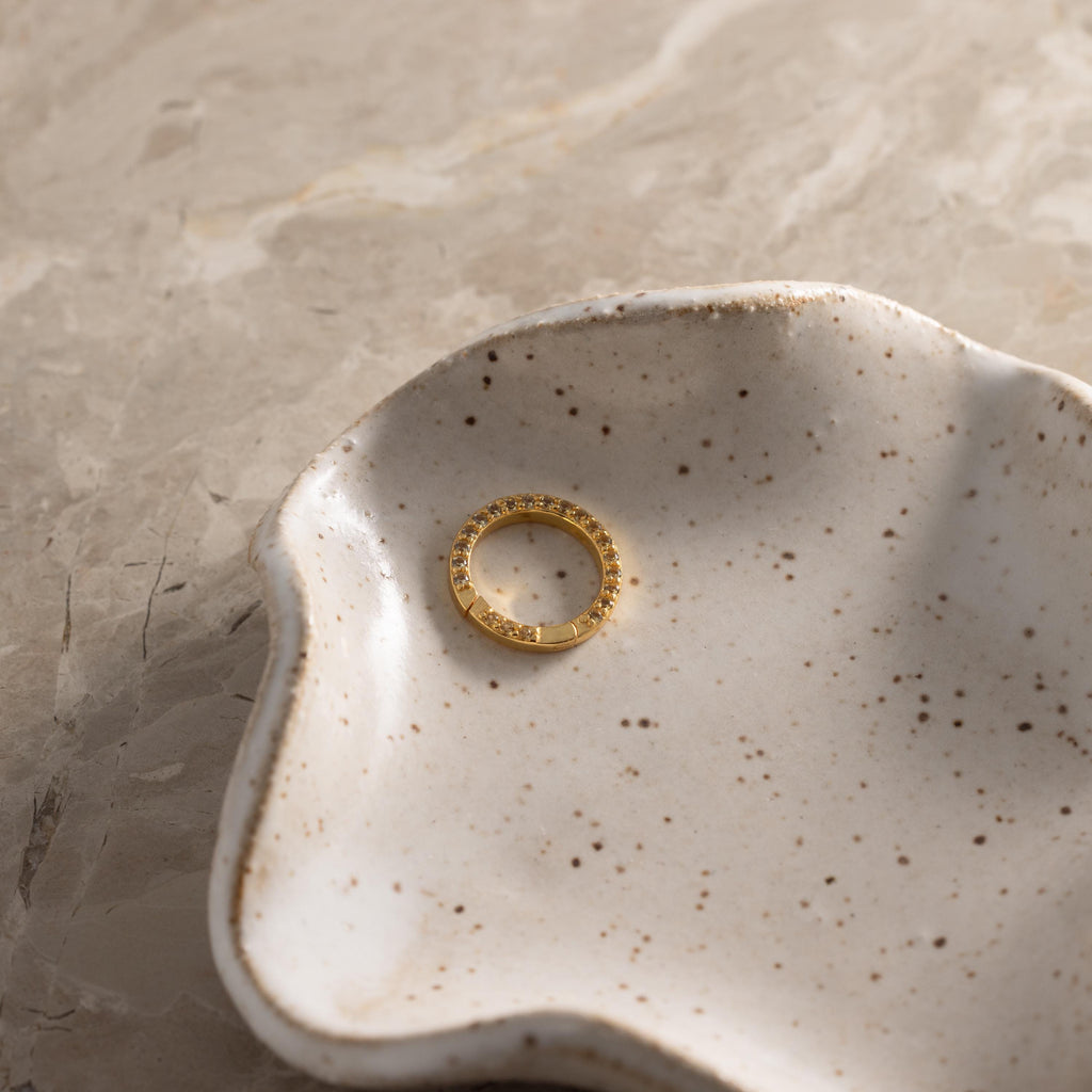 A Pave Topaz Charm Clip sits in a speckled, wavy-edged ceramic dish atop a beige marble surface.