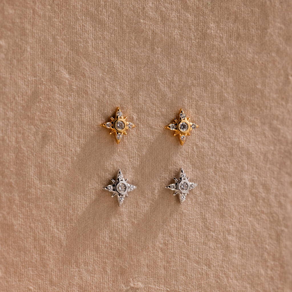 One pair of gold Pave North Star Earrings and one pair of silver Pave North Star Earrings displayed on a tan surface