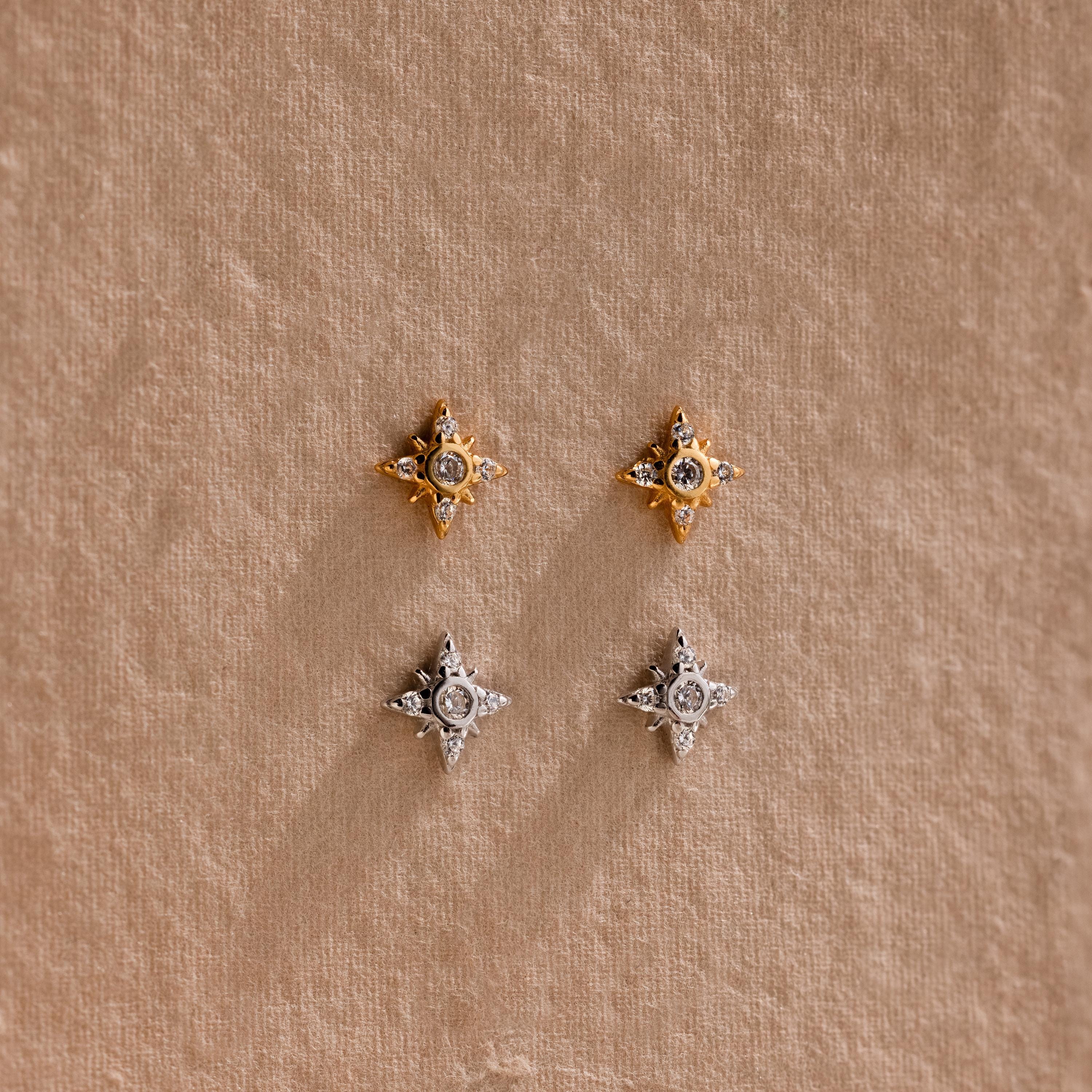 One pair of gold Pave North Star Earrings and one pair of silver Pave North Star Earrings displayed on a tan surface