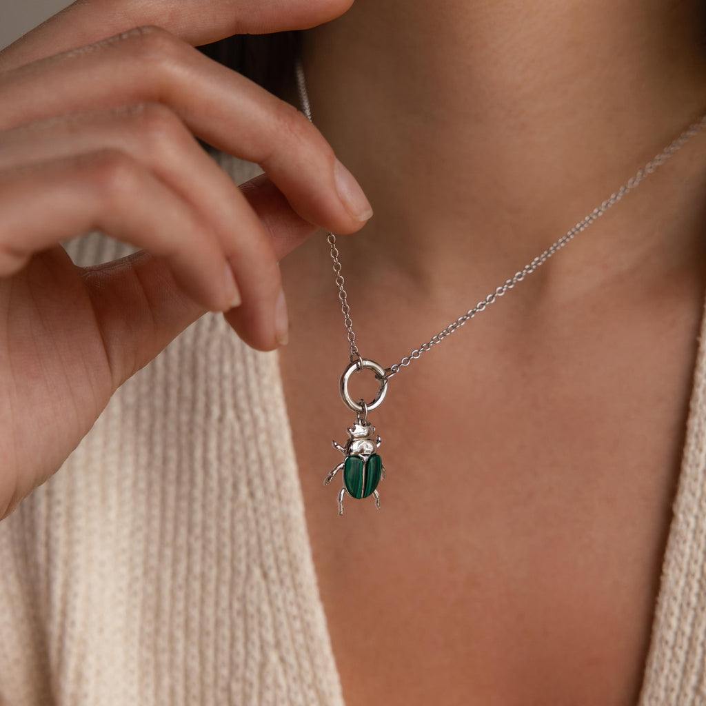 A silver chain necklace with the silver version of the beetle charm is worn around the neck of a model, shown against a cream knit top.