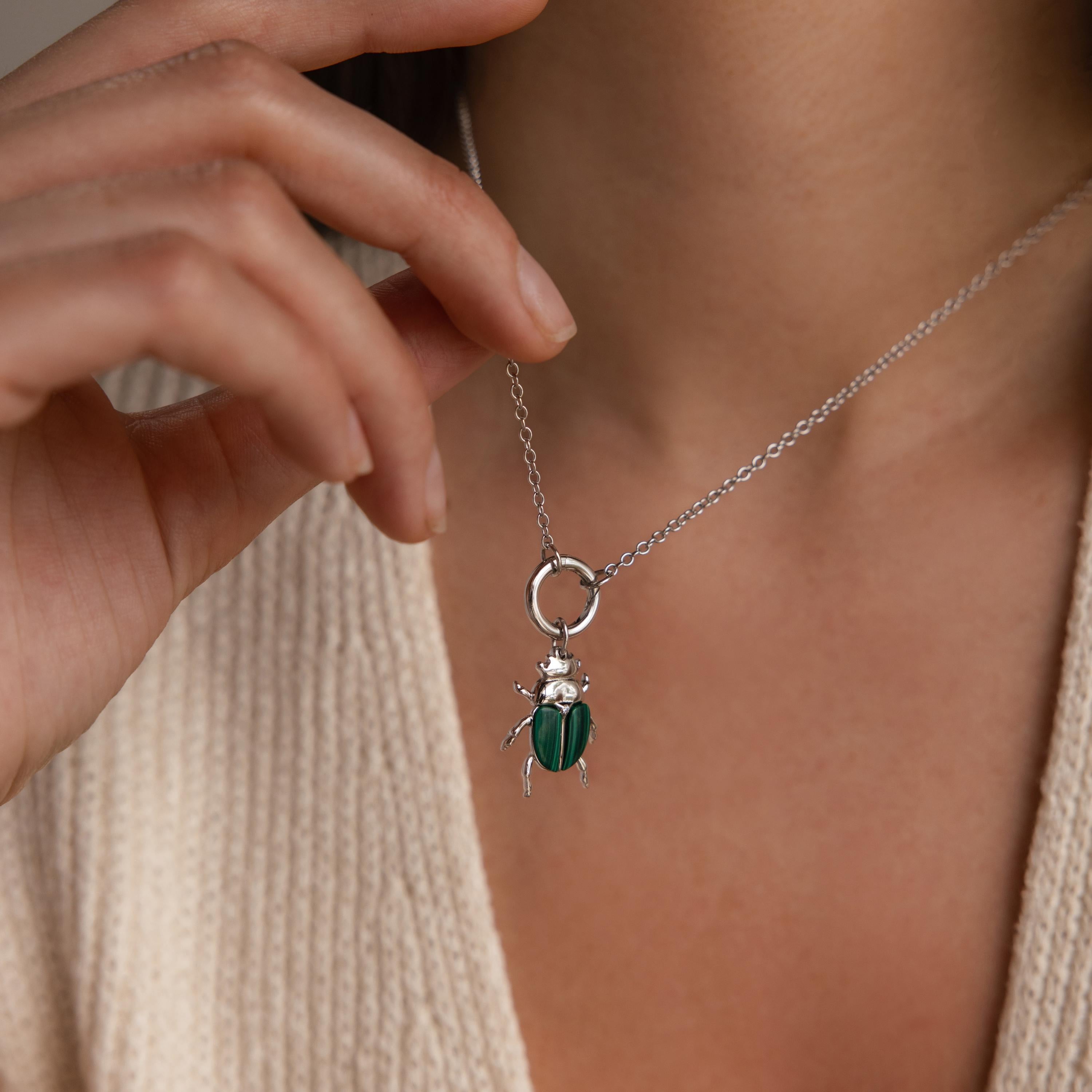 A silver chain necklace with the silver version of the beetle charm is worn around the neck of a model, shown against a cream knit top.