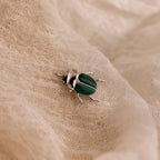 A silver beetle charm with malachite green wings rests on a soft, textured cream fabric.