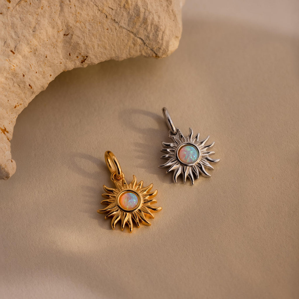 Two Opal Sun Charms, each with a luminous opal center—one in gold and one in silver—are displayed on a beige surface.
