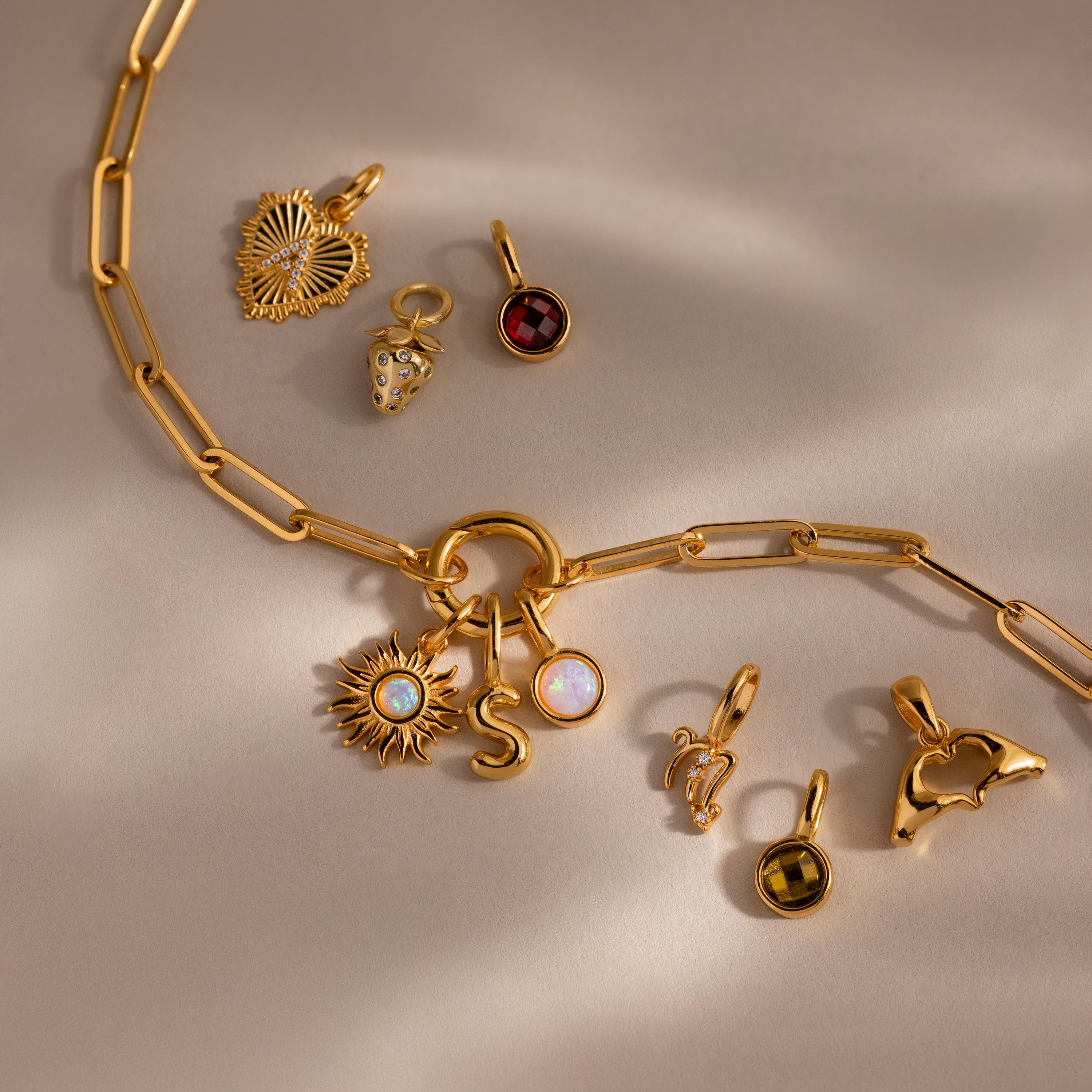 The Opal Sun Charm features gold chains adorned with initials, gemstones, a golden sun, heart, and zodiac charms—plus a luminous opal accent—all set against a beige background.