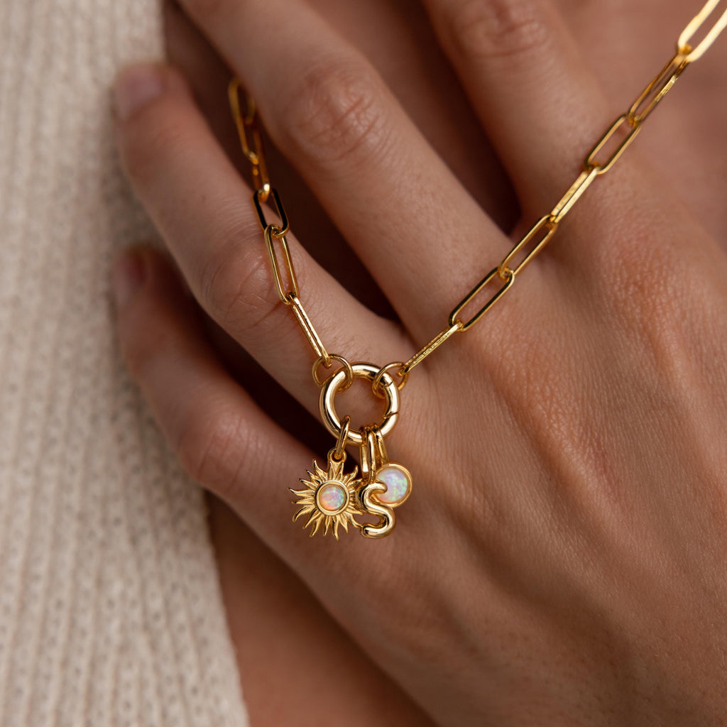 The Opal Sun Charm is shown on a gold necklace with two additional charms: a small round opal disc and an “S” initial, all hanging from a chunky round clasp held against the back of a hand.