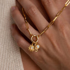 The Opal Sun Charm is shown on a gold necklace with two additional charms: a small round opal disc and an “S” initial, all hanging from a chunky round clasp held against the back of a hand.