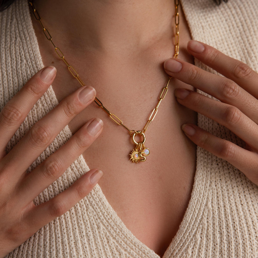 A woman wears a gold chain necklace with the Opal Sun Charm, opal disc charm, and an “S” initial charm centered on the chest, styled with a deep V-neck knit top.