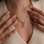 A woman wears a gold chain necklace with the Opal Sun Charm, opal disc charm, and an “S” initial charm centered on the chest, styled with a deep V-neck knit top.