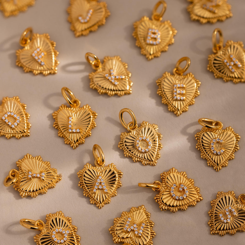 A scattered flat lay of Ribbed Heart Initial Pendants featuring various letters such as L O G K E B and more, all displayed with radiant gold finishes on a soft beige backdrop.