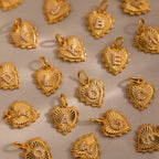 A scattered flat lay of Ribbed Heart Initial Pendants featuring various letters such as L O G K E B and more, all displayed with radiant gold finishes on a soft beige backdrop.