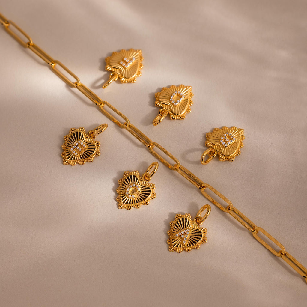 Six Ribbed Heart Initial Pendants with letters E C A B D F  arranged beside a chunky paperclip chain on a neutral surface, showing personalization options.