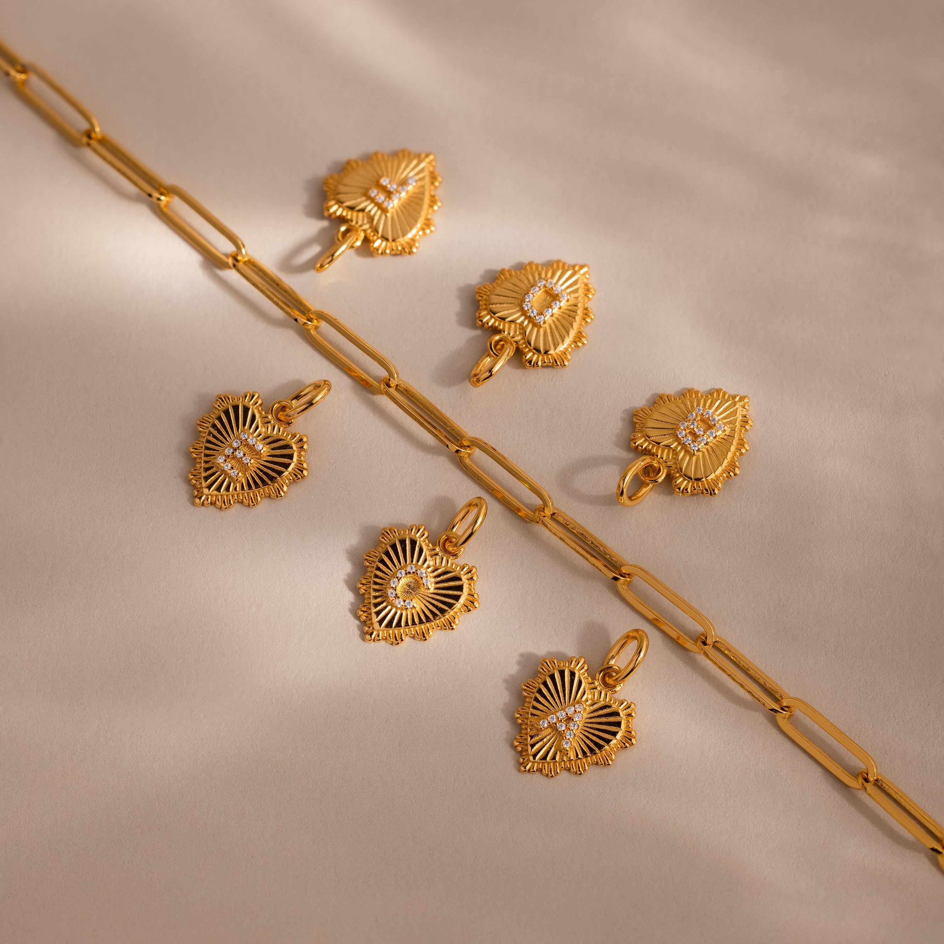 Six Ribbed Heart Initial Pendants with letters E C A B D F  arranged beside a chunky paperclip chain on a neutral surface, showing personalization options.