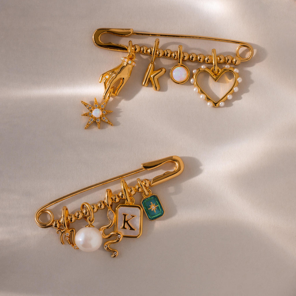 Two fully styled beaded safety pin charms laid flat—one features a celestial hand, opal star, initial K, and heart charm, while the other includes a pearl, snake, and green enamel charm.