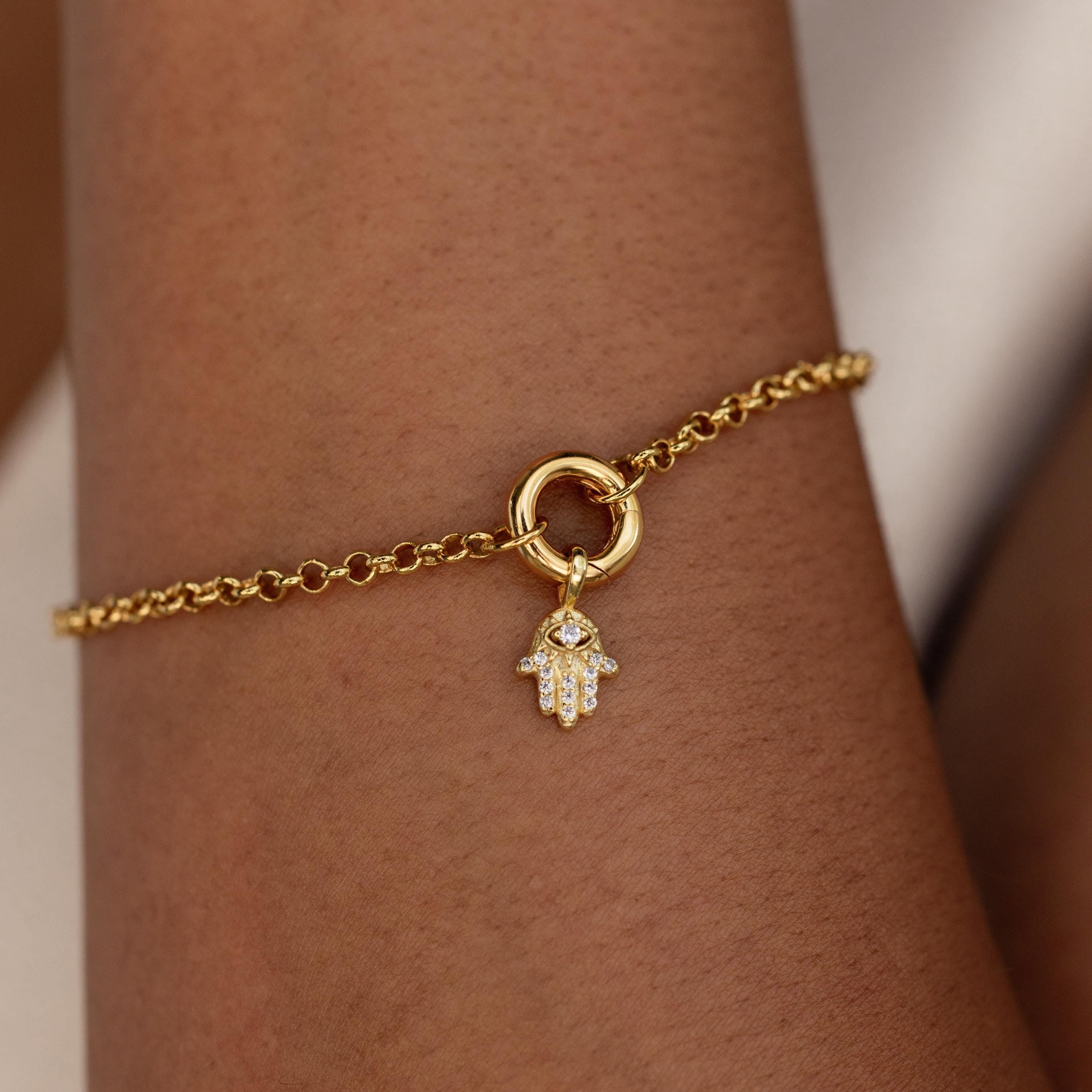A gold bracelet worn on the wrist featuring a dainty Evil Eye Hand Charm dangling from a round clasp.
