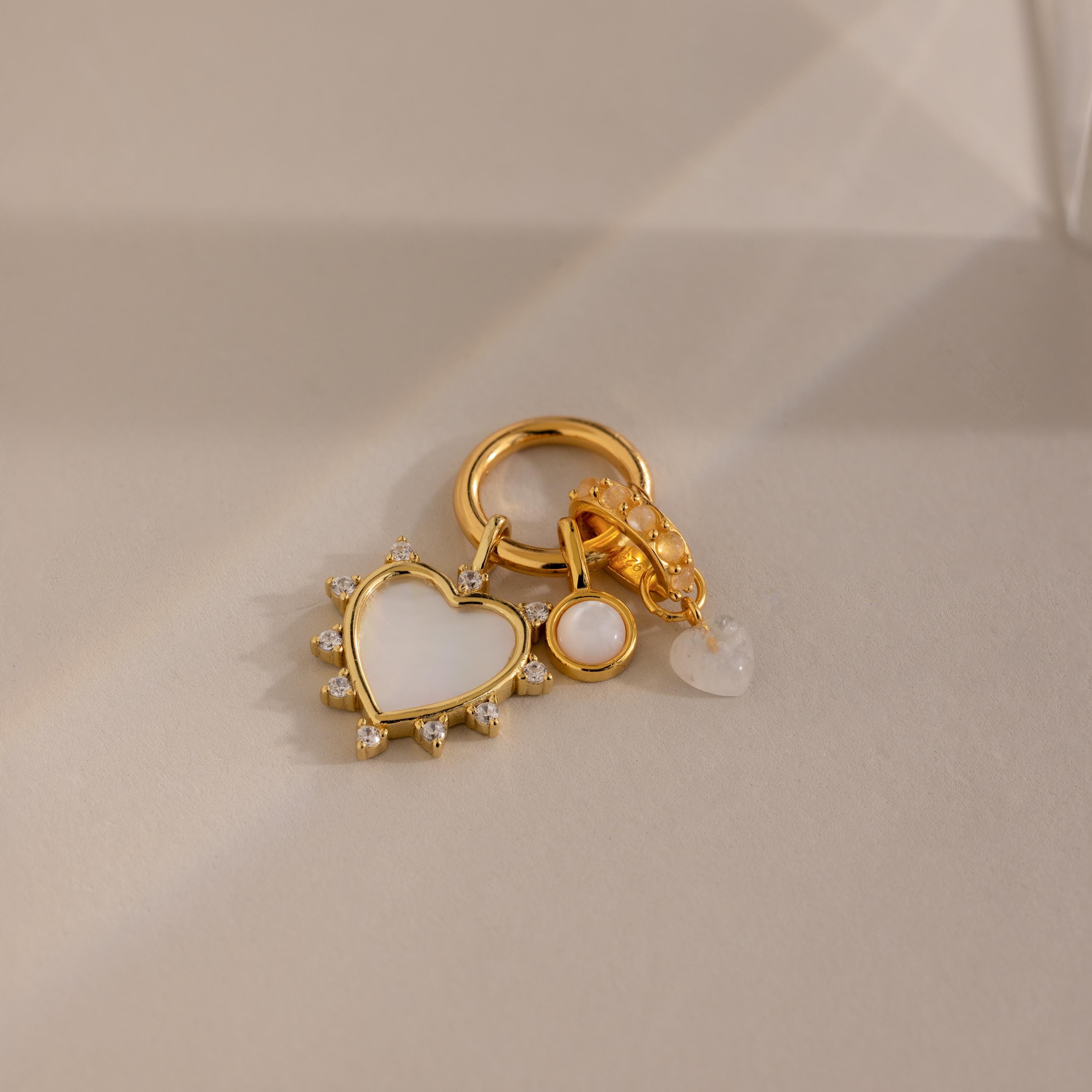 The gold charm is styled on a round connector clasp alongside two other charms: a pink mother-of-pearl disc and a small translucent heart.