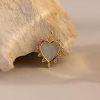 A single gold Starburst Pearl Heart Charm is displayed leaning against a beige stone, glowing under natural light.